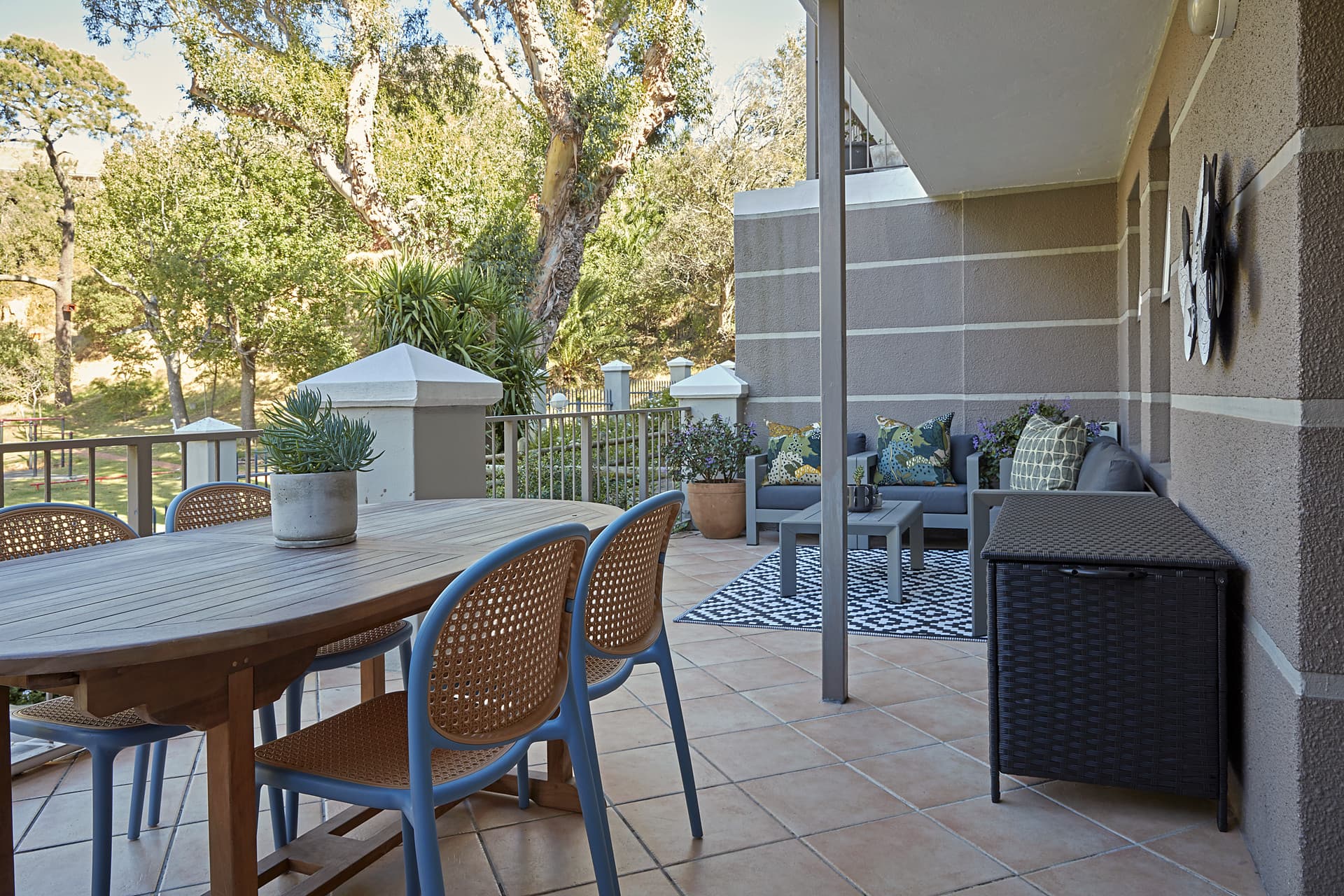 Gardens – Covered balcony with a timber dining table, cane-backed chairs and a seating corner, looking out into the trees