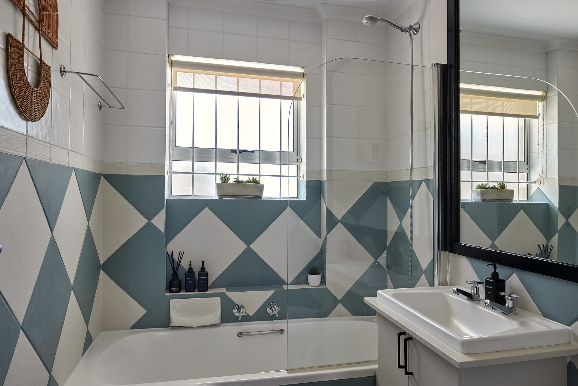Gardens – Bathroom with the bath under the window, a diamond-patterned teal and cream tiled dado and a black-framed mirror over the basin