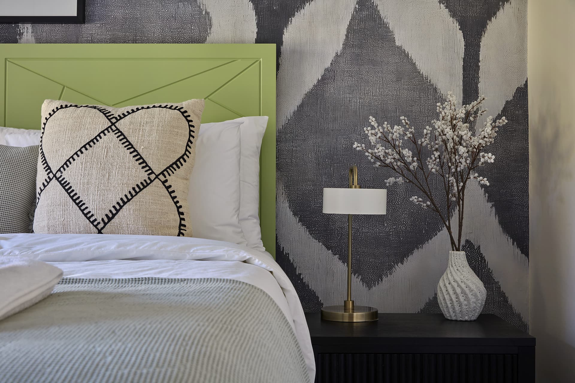 Gardens – Bedside table with a brass lamp and a textured white vase of blossom against the ikat wall