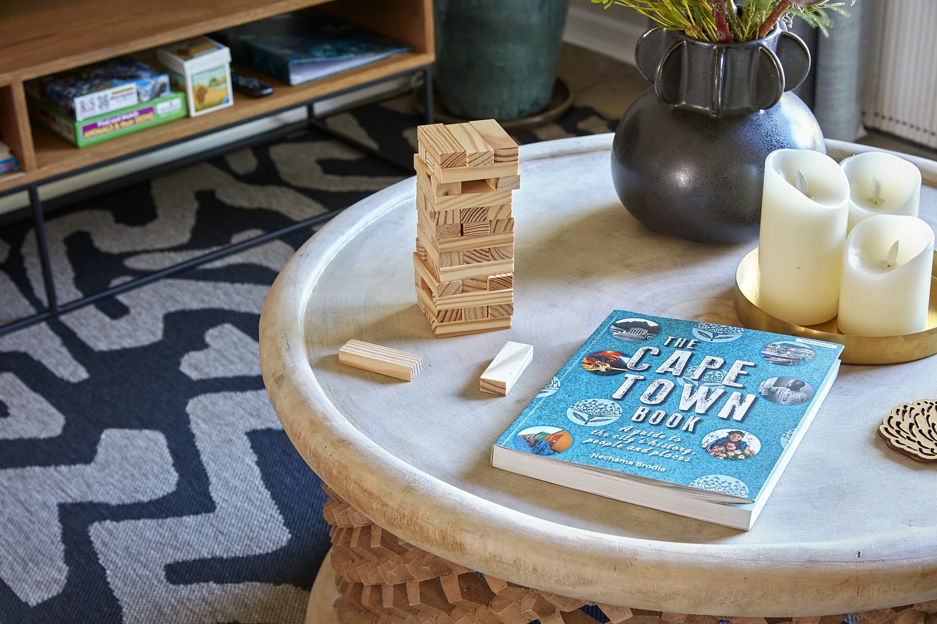 Gardens – Round pale timber coffee table with a stack of wooden blocks, pillar candles on a brass tray and a Cape Town guidebook