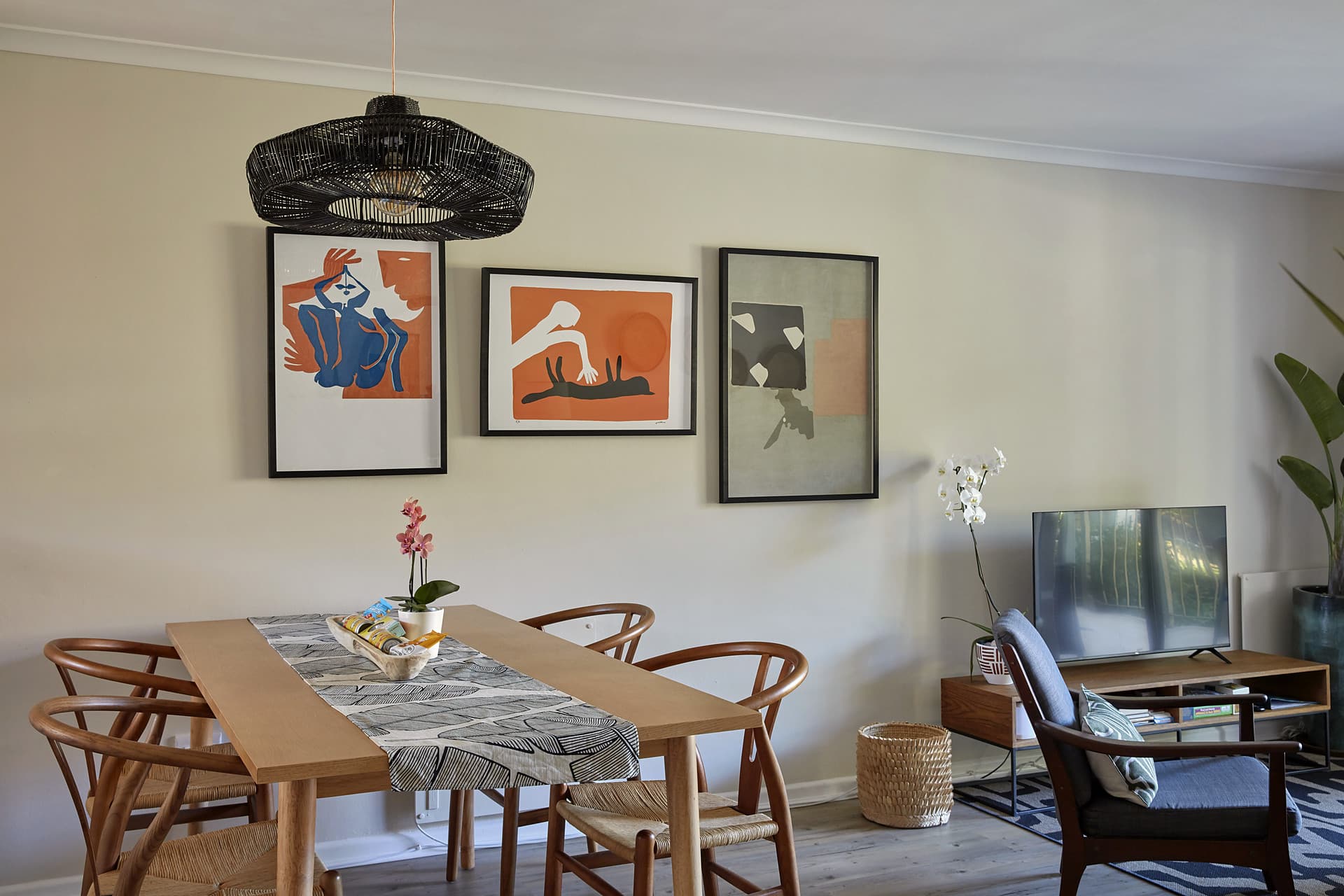 Gardens – Dining table and wishbone chairs beneath three framed prints and a black woven pendant