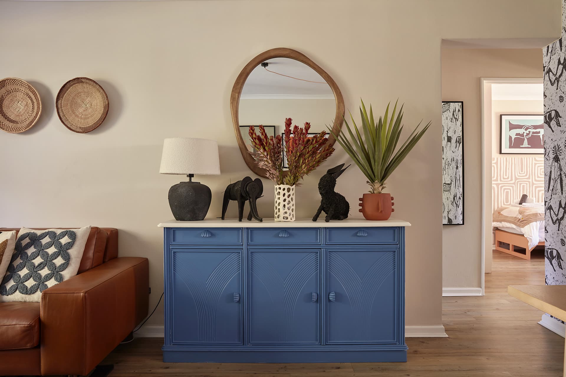 Gardens – Blue fluted sideboard beneath a round timber mirror, with a black table lamp, carved animal figures and a planted aloe