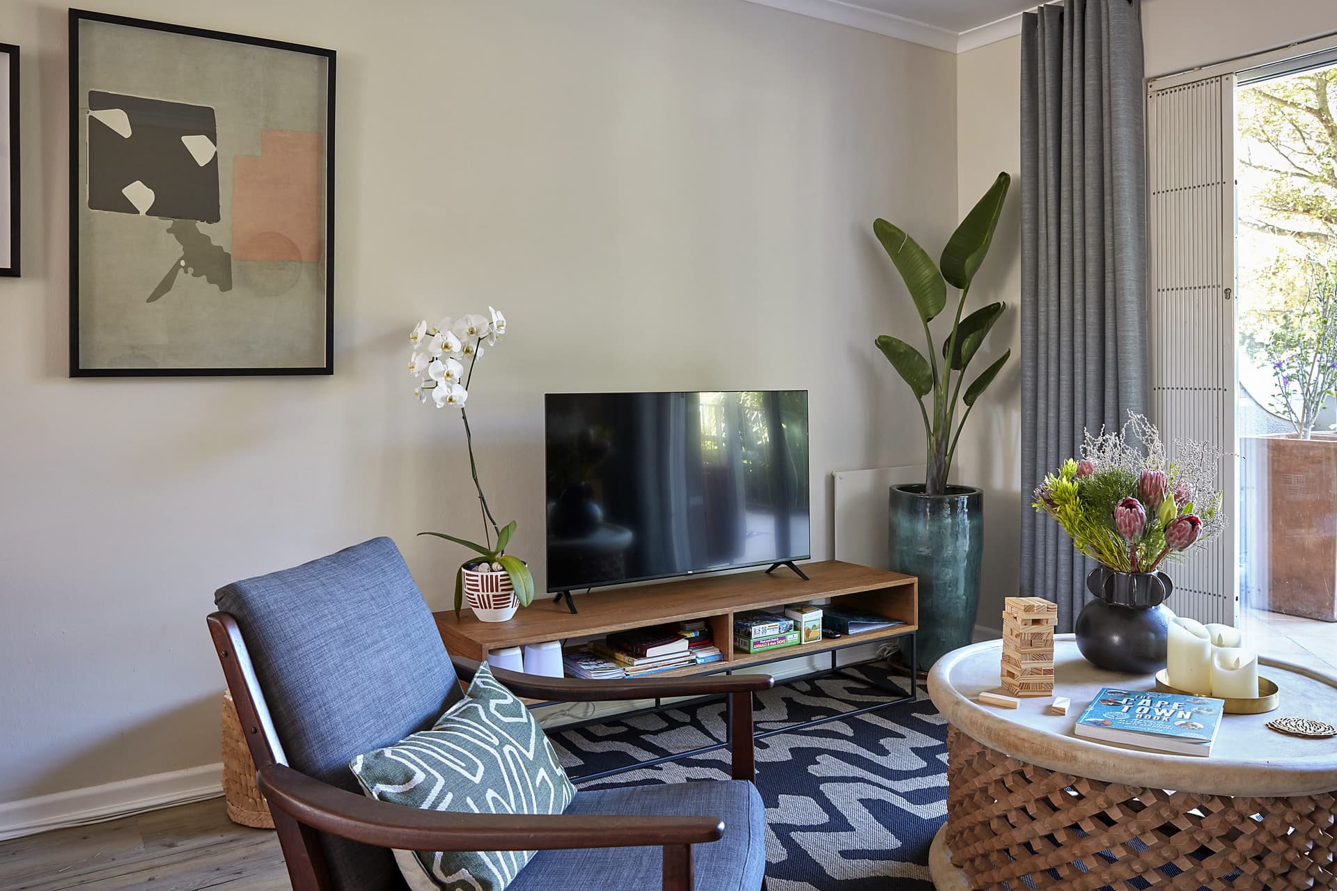 Gardens – Living corner with a blue upholstered armchair, a low oak media unit, a potted strelitzia and curtains to the balcony