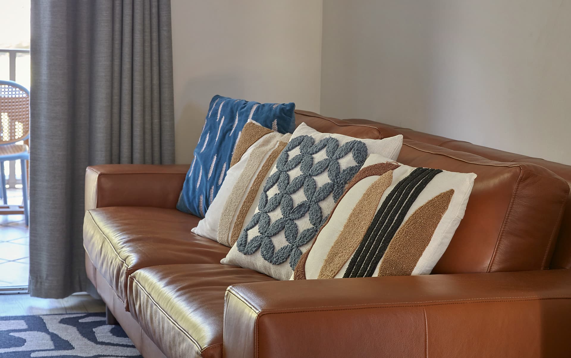 Gardens – Tan leather sofa with blue, cream and textured woven cushions beside a curtained window