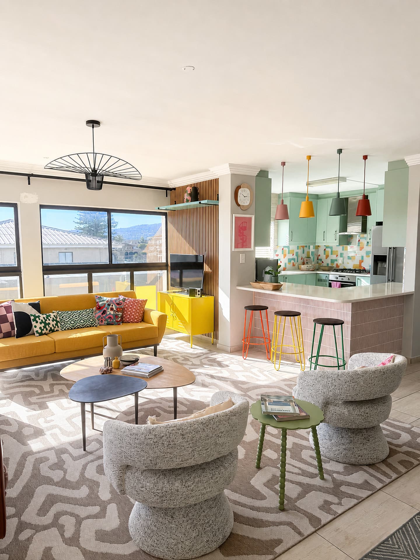 Strand – Living room toward the kitchen, sunlight on the rug, yellow sofa and pink tiled island