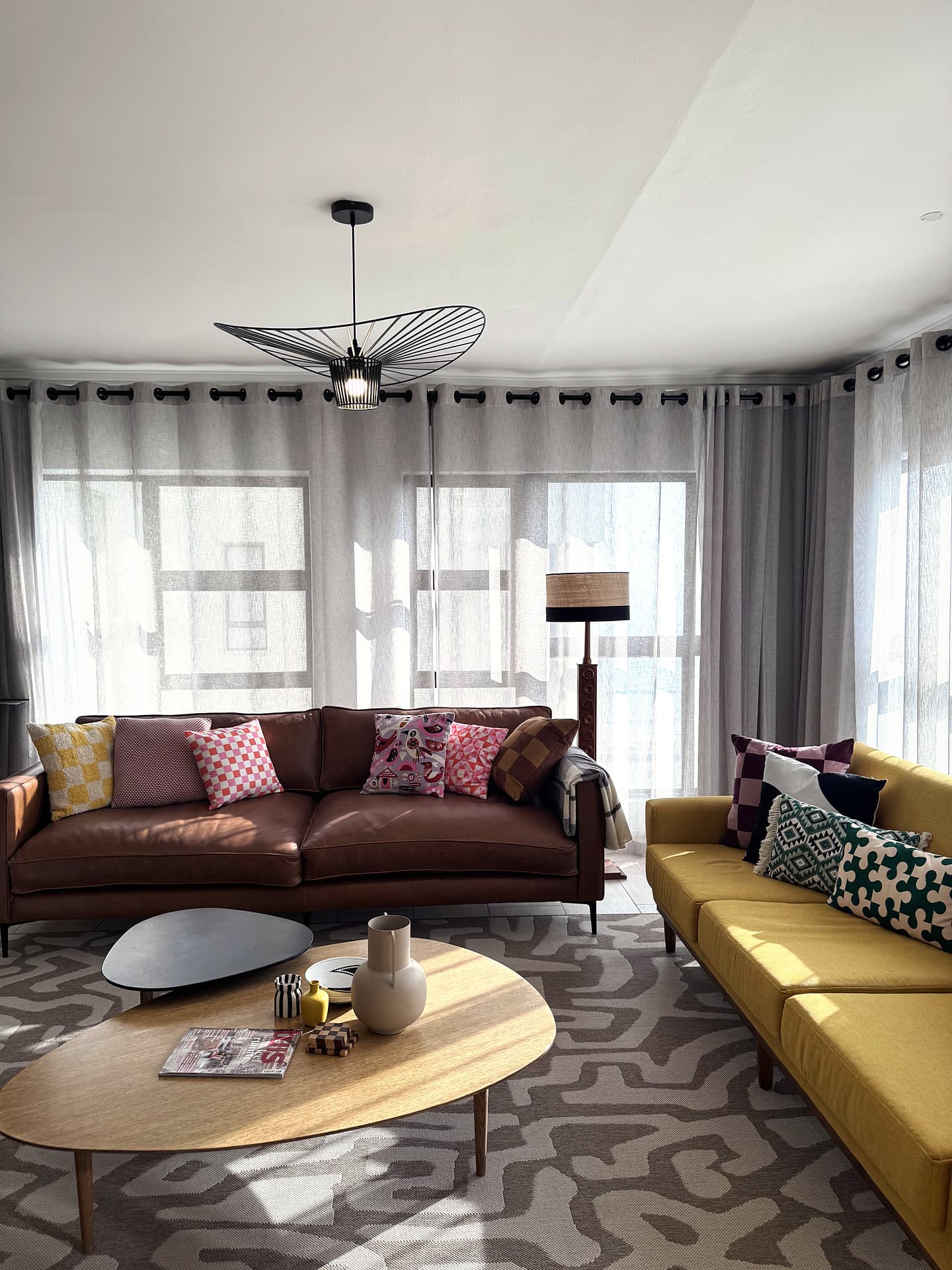 Strand – Living room with a brown leather sofa, yellow sofa, patterned rug and a wire pendant, sheer curtains behind