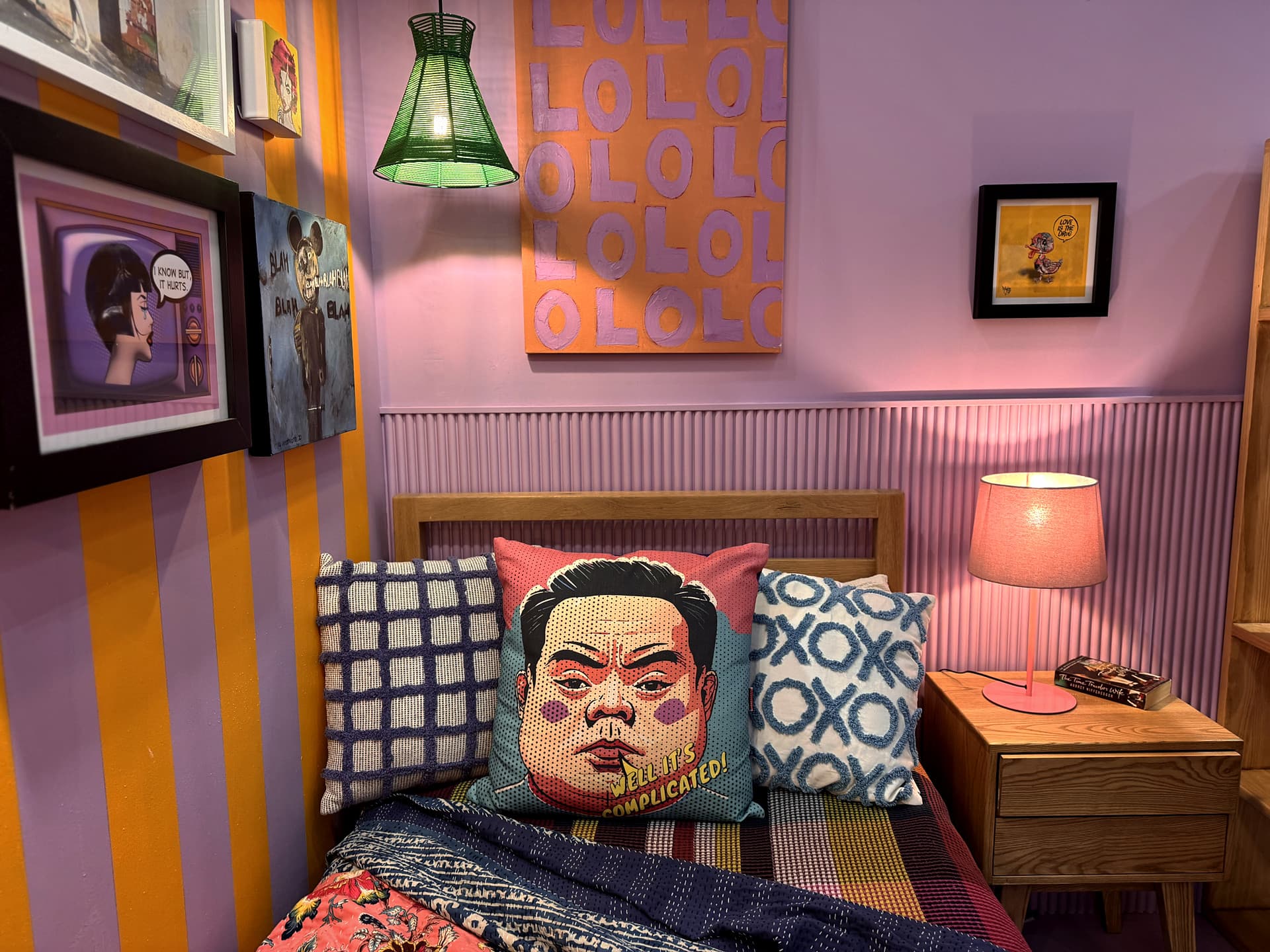 Urban Living Pod – Bed corner with patterned cushions, an art wall of small canvases and a green pendant